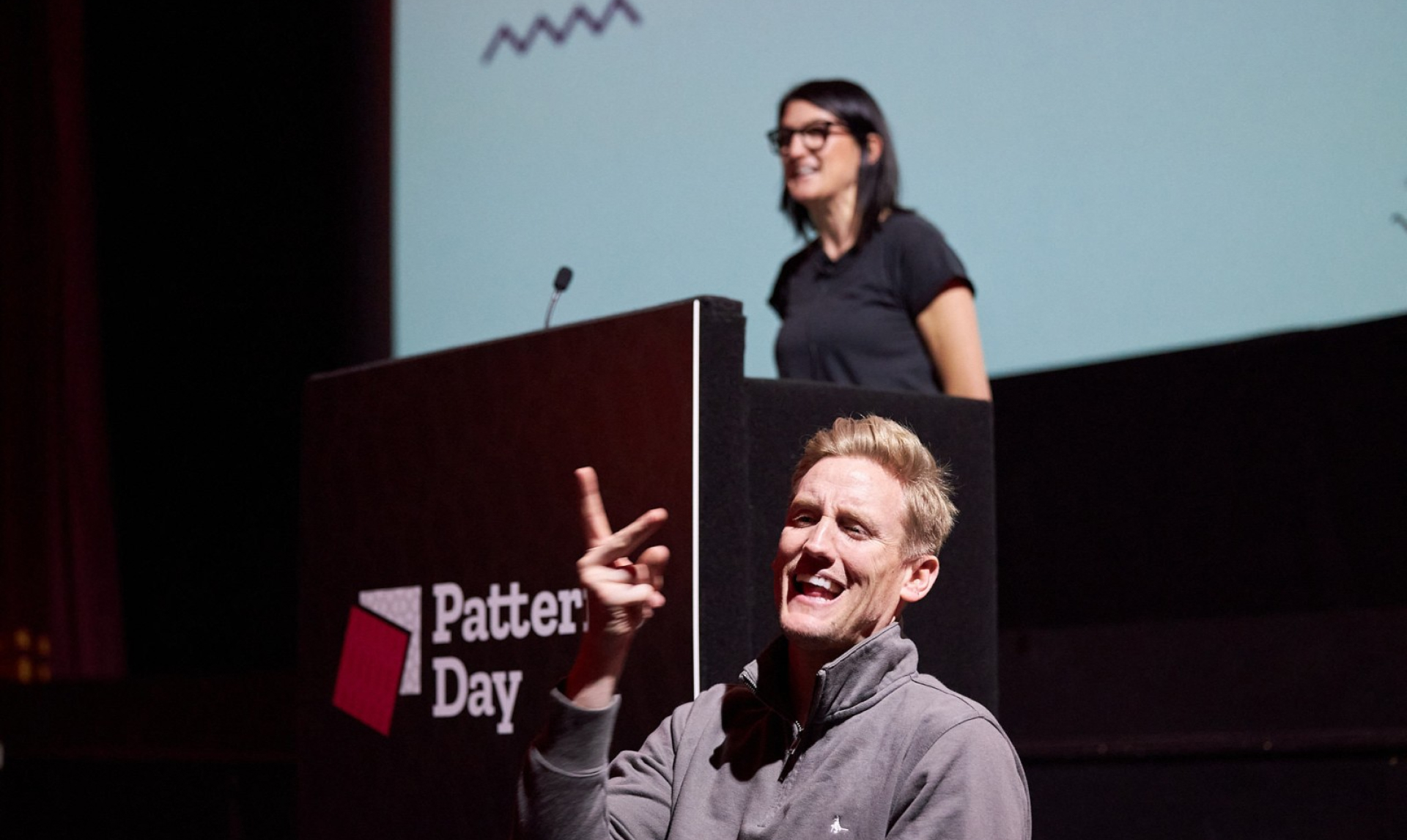 Geri Reid speaking on stage at Patterns Day with a British Sign Language interpreter signing at the front of the stage
